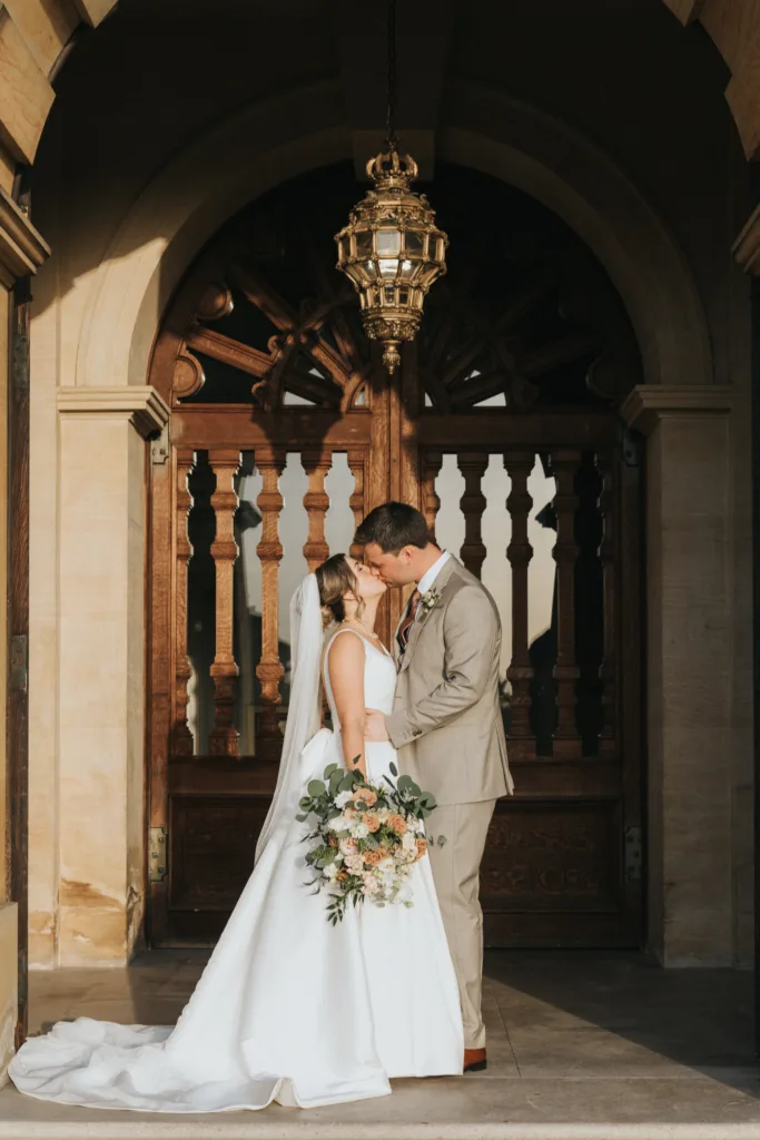 A bride and groom stand close, touching noses and smiling, in front of ornate wooden doors and arched stonework at their Harlaxton Manor wedding. The bride wears a white gown and veil, holding a bouquet of white and peach flowers; the groom a light tan suit. © Aimee Lince Photography, Lincolnshire photographer