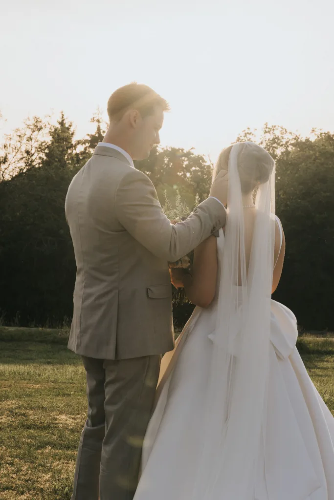 A groom in a light grey suit gently touches the bride’s face as they stand outdoors at sunset during their Harlaxton Manor Wedding. The bride wears a white dress with a long veil and large bow, both facing away amid greenery and soft sunlight. © Aimee Lince Photography, Lincolnshire photographer