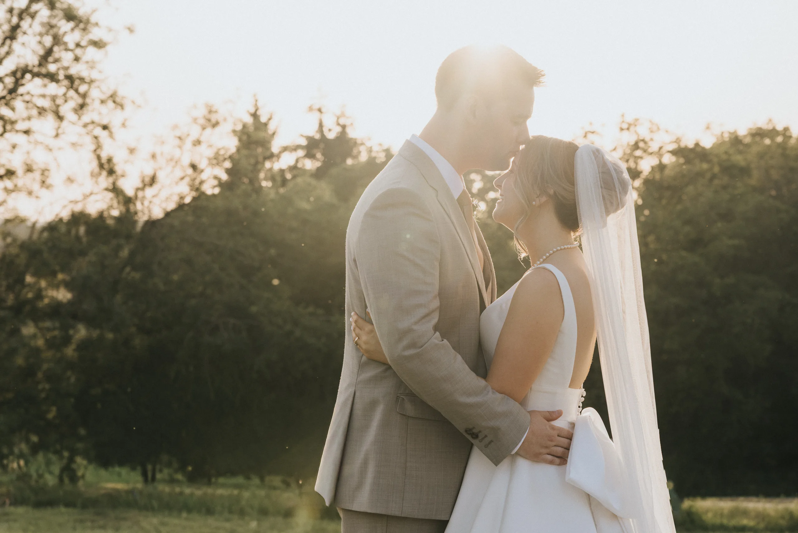 A bride in a white dress and veil embraces her groom in a tan suit at sunset during their Harlaxton Manor wedding, sharing a forehead-to-forehead moment outdoors as sunlight creates a warm, romantic glow among the trees. © Aimee Lince Photography, Lincolnshire photographer