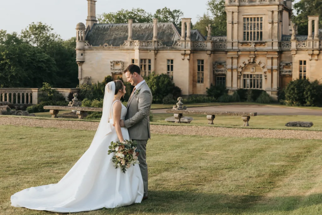 A bride in a white dress and veil stands facing her groom on a manicured lawn at their Harlaxton Manor wedding, holding a bouquet. They gaze at each other affectionately, with the ornate historic mansion and lush greenery behind them. © Aimee Lince Photography, Lincolnshire photographer
