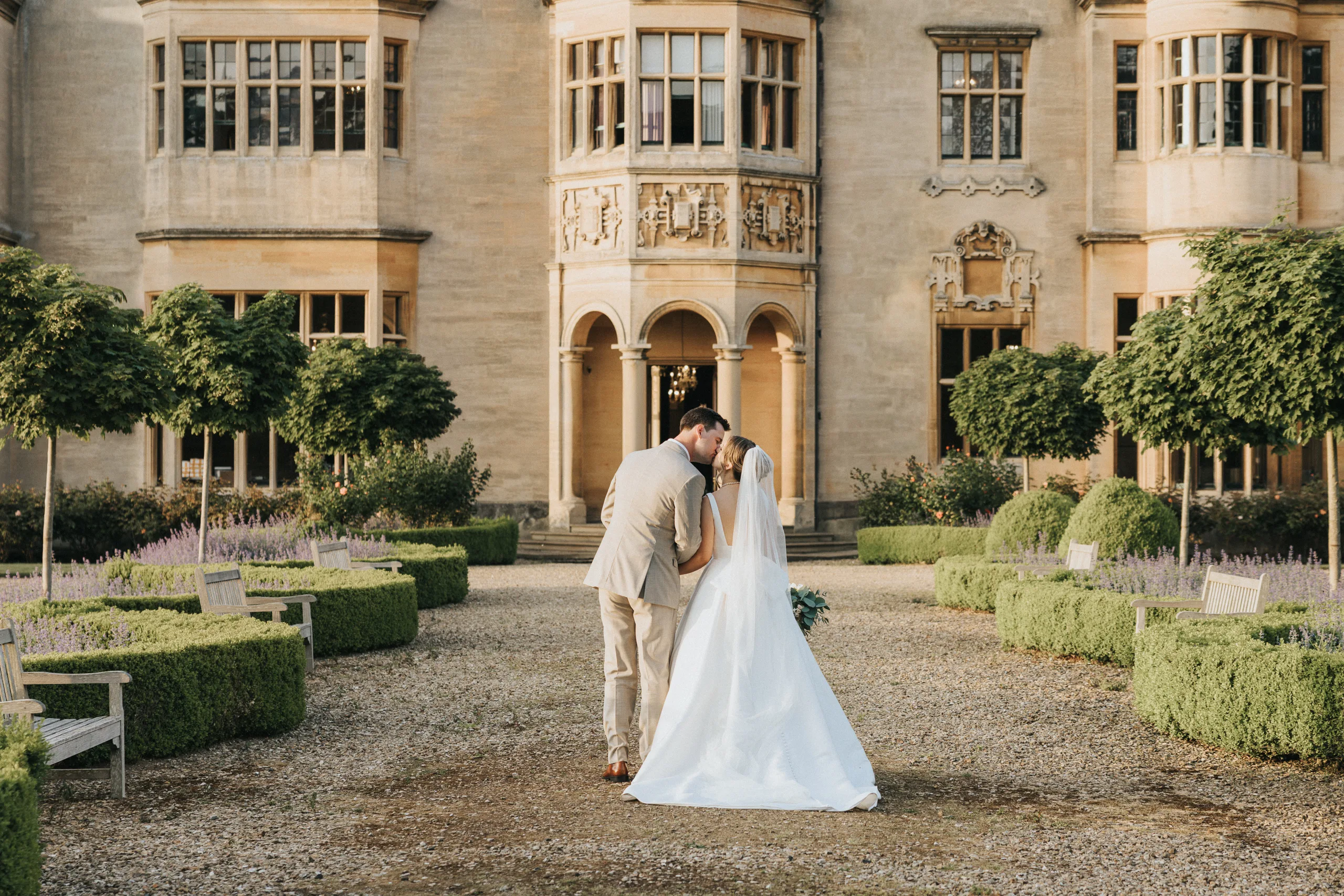 A bride and groom in wedding attire walk hand-in-hand down a garden path at their Harlaxton Manor Wedding, surrounded by manicured hedges and trees, towards a grand historic stone building with large windows and arched entryways. © Aimee Lince Photography, Lincolnshire photographer