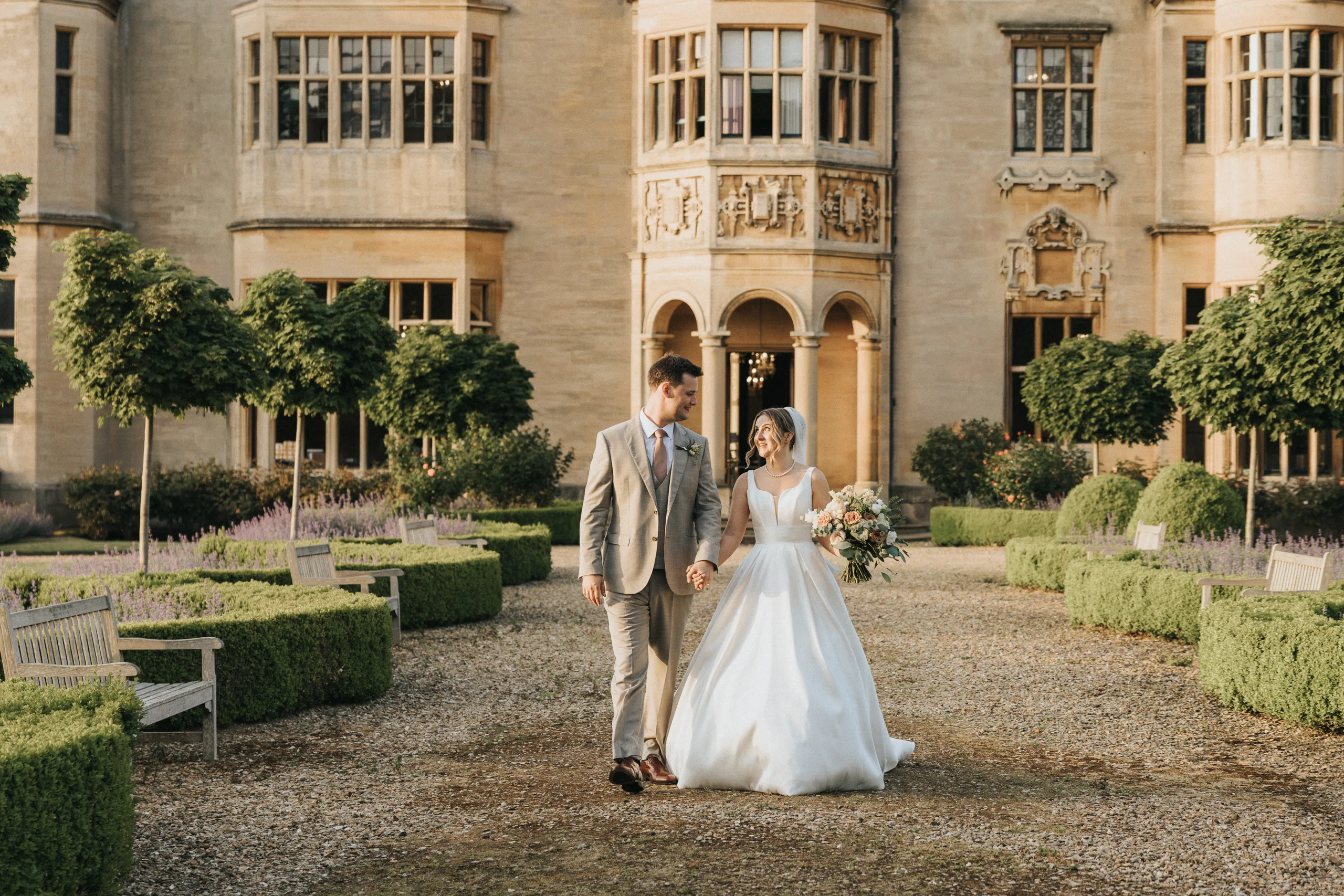 A bride and groom walk hand in hand outside grand Harlaxton Manor, its large windows towering above. The bride’s white gown flows as they stroll the gravel path, lined with green hedges and benches, soaking in their enchanting Harlaxton Manor wedding gardens. © Aimee Lince Photography, Lincolnshire photographer