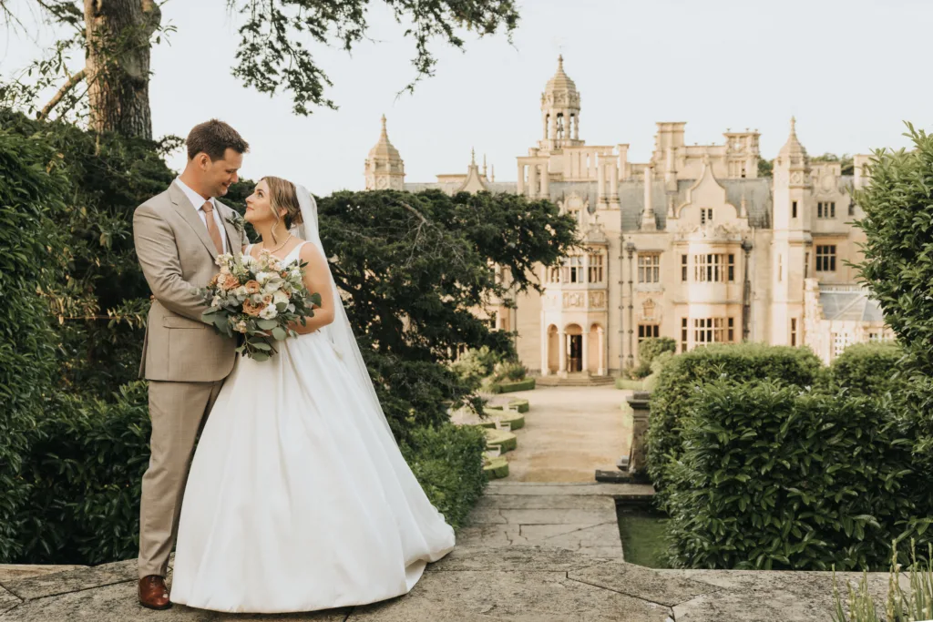 A bride and groom stand facing each other, smiling in elegant wedding attire. The bride holds a large bouquet. Behind them, Harlaxton Manor’s historic stone towers and lush greenery create a romantic, fairy-tale wedding atmosphere. © Aimee Lince Photography, Lincolnshire photographer
