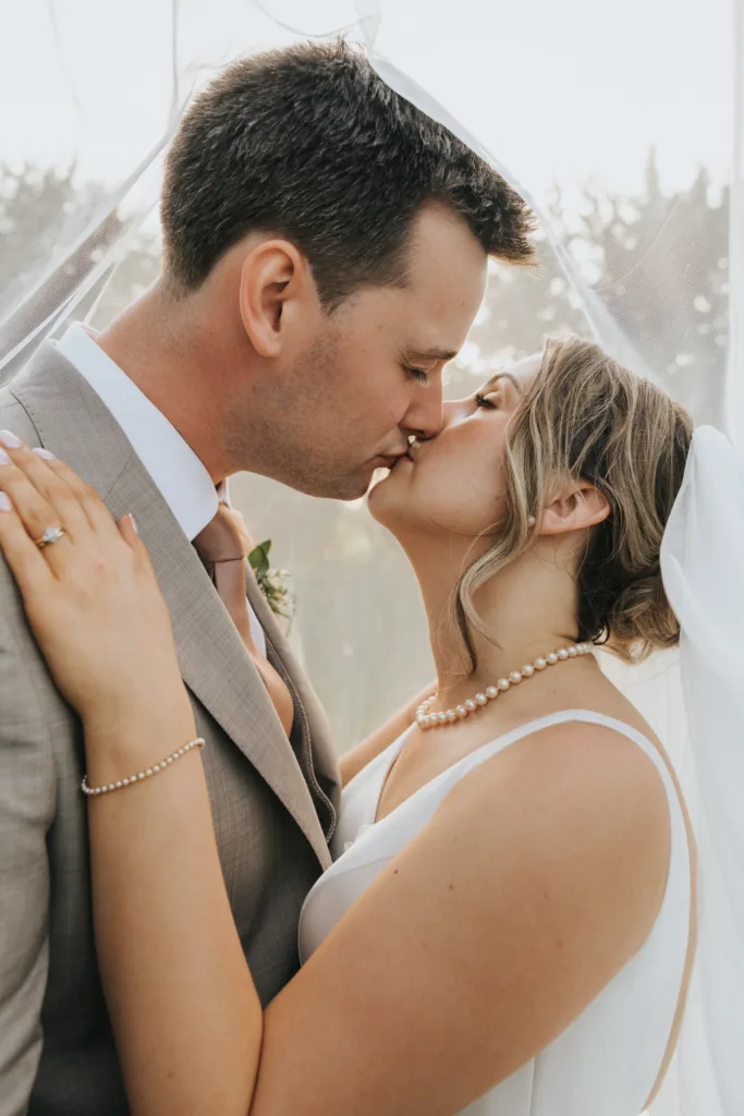 A bride and groom share a gentle kiss under a sheer veil at their Harlaxton Manor wedding. The groom wears a light grey suit, the bride a white dress and pearl necklace. Her hand, adorned with a wedding ring, rests lovingly on his shoulder. © Aimee Lince Photography, Lincolnshire photographer