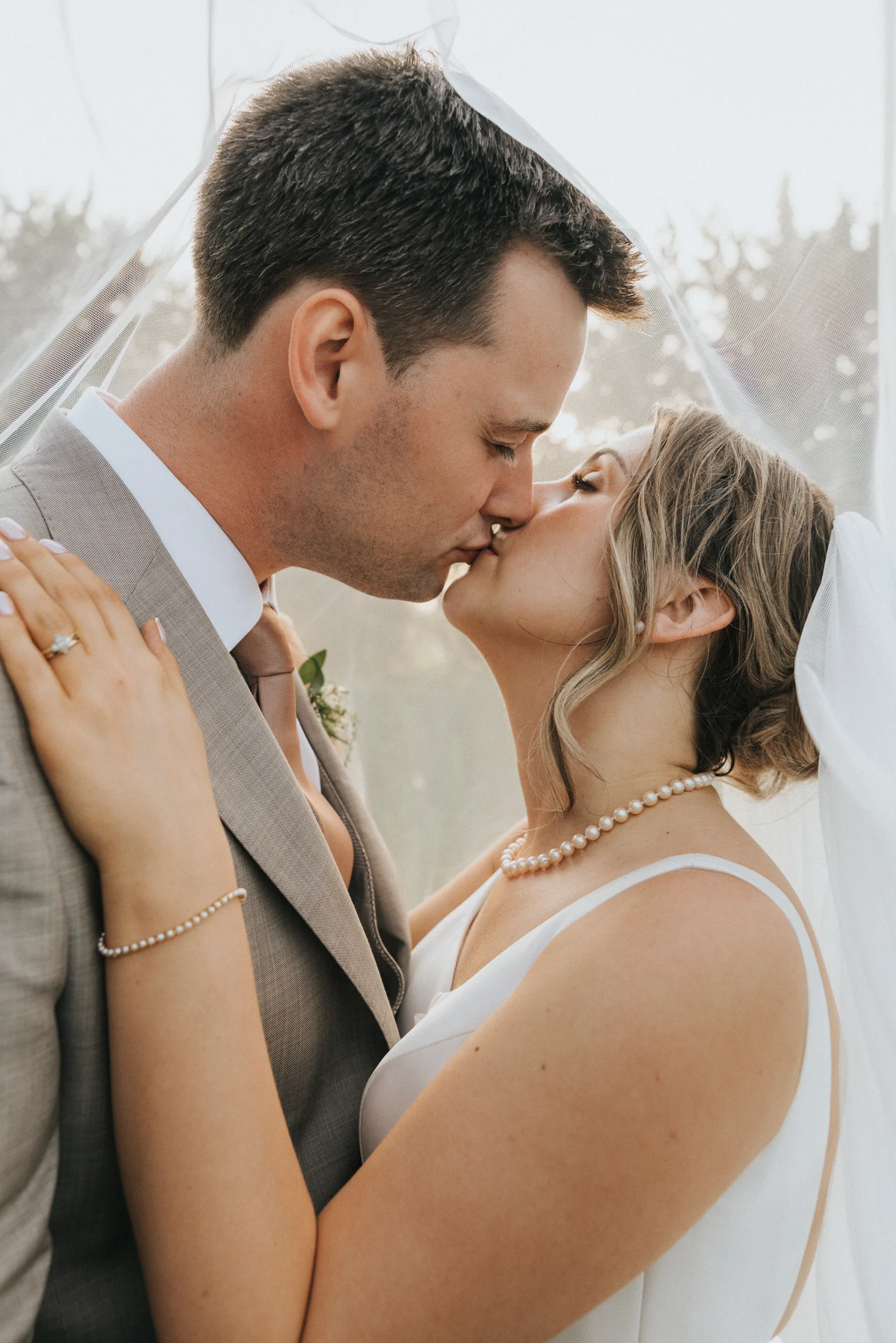 A bride and groom share a gentle kiss under a sheer veil at their Harlaxton Manor wedding. The groom wears a light grey suit, the bride a white dress and pearl necklace. Her hand, adorned with a wedding ring, rests lovingly on his shoulder. © Aimee Lince Photography, Lincolnshire photographer