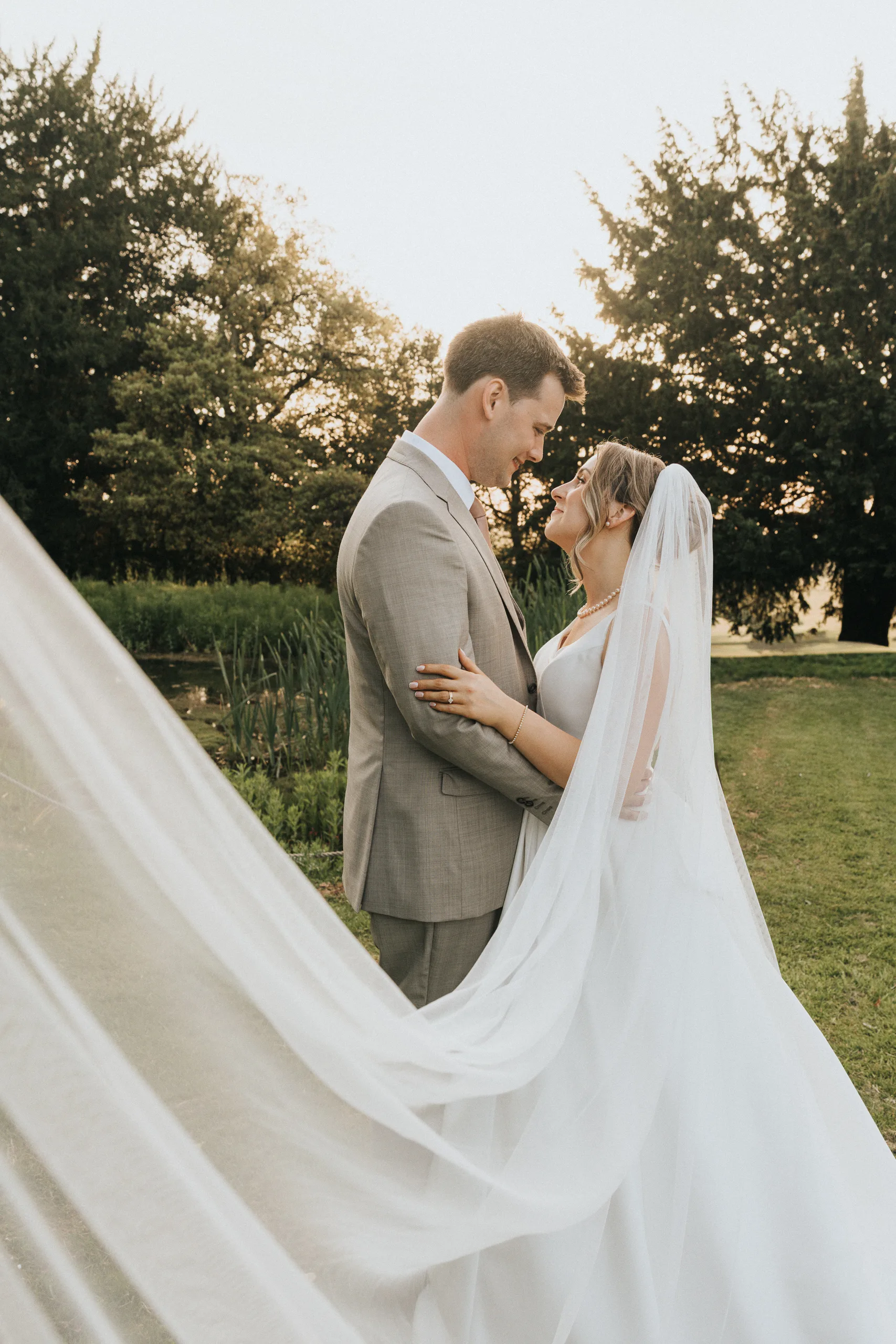 A bride in a white gown and veil stands close to a groom in a light grey suit. They embrace outdoors at their Harlaxton Manor wedding, gazing into each other's eyes as sunlight filters through the trees and her veil flows softly in the foreground. © Aimee Lince Photography, Lincolnshire photographer