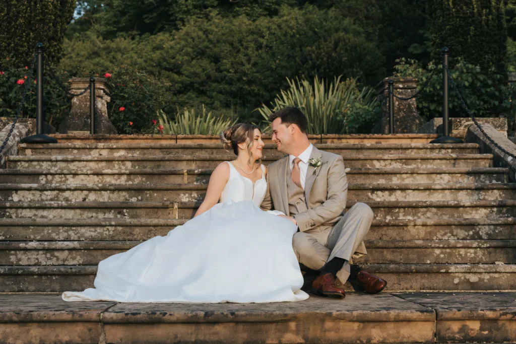 A bride in a white dress and a groom in a beige suit sit close together on stone steps outdoors at their Harlaxton Manor Wedding, touching foreheads and smiling lovingly, with lush greenery and flowers in the background. © Aimee Lince Photography, Lincolnshire photographer