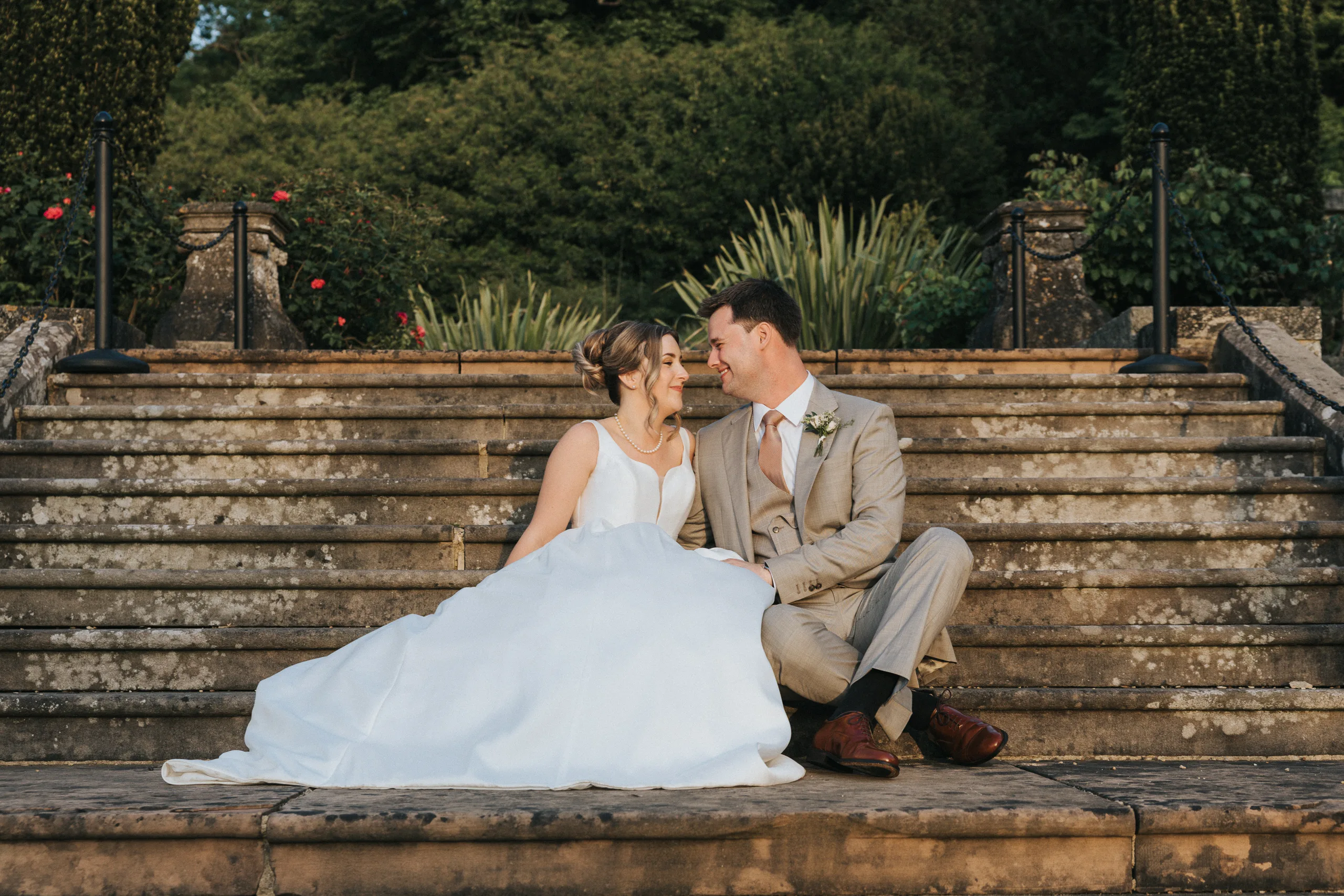 A bride in a white dress and a groom in a beige suit sit close together on stone steps outdoors at their Harlaxton Manor Wedding, touching foreheads and smiling lovingly, with lush greenery and flowers in the background. © Aimee Lince Photography, Lincolnshire photographer