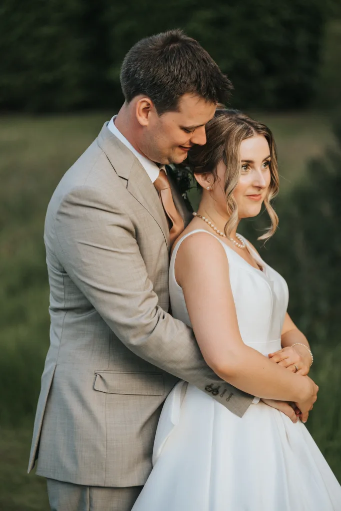 A groom in a light beige suit gently embraces a bride in a white dress with pearl jewellery. They stand outdoors amid lush green foliage at their Harlaxton Manor wedding. The bride faces forward, smiling softly, while the groom looks at her affectionately. © Aimee Lince Photography, Lincolnshire photographer