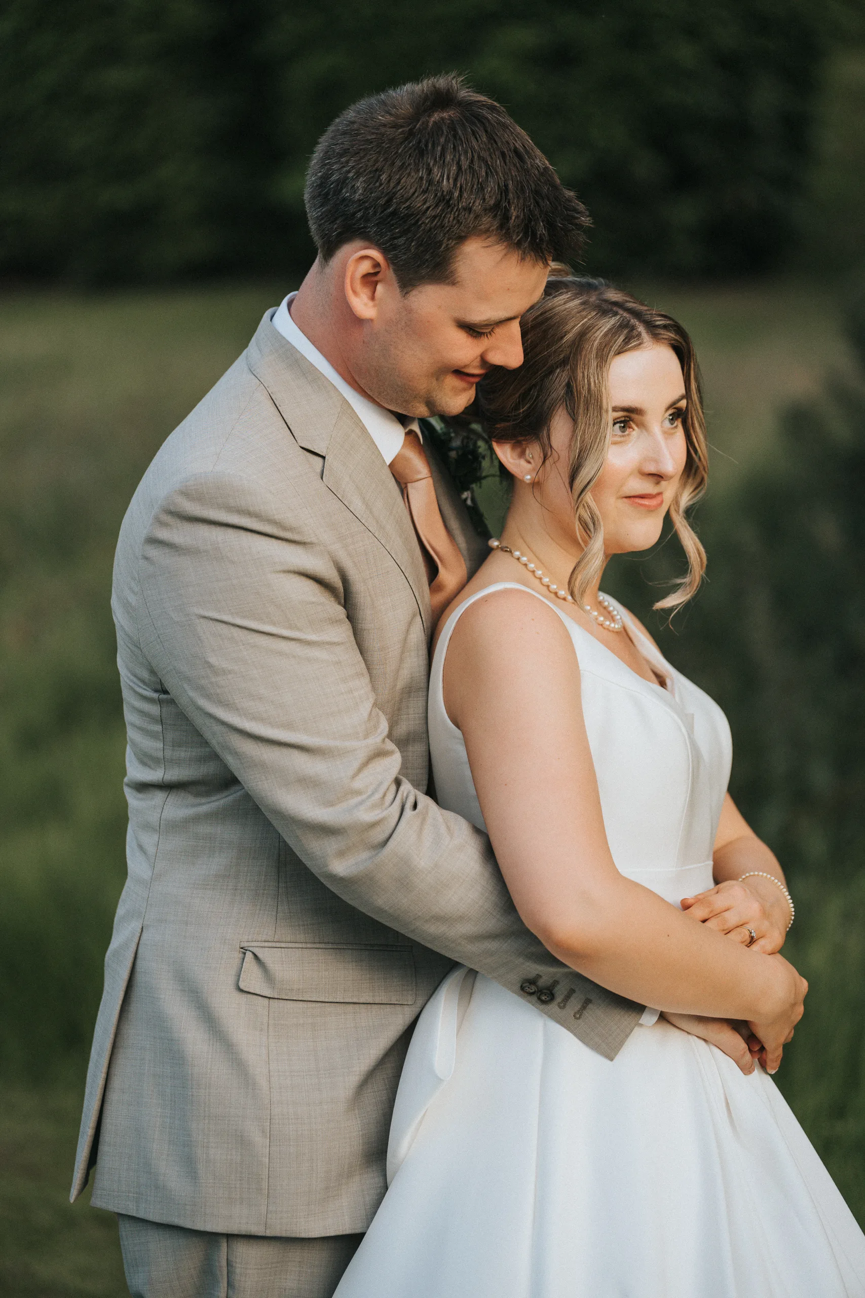 A groom in a light beige suit gently embraces a bride in a white dress with pearl jewellery. They stand outdoors amid lush green foliage at their Harlaxton Manor wedding. The bride faces forward, smiling softly, while the groom looks at her affectionately. © Aimee Lince Photography, Lincolnshire photographer