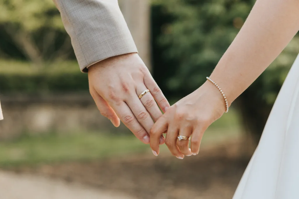Close-up of two people holding hands outdoors at a Harlaxton Manor Wedding. Both wear rings on their ring fingers—one in a beige suit, the other in a white dress with a delicate bracelet. Blurred greenery and a stone wall complete the scene. © Aimee Lince Photography, Lincolnshire photographer