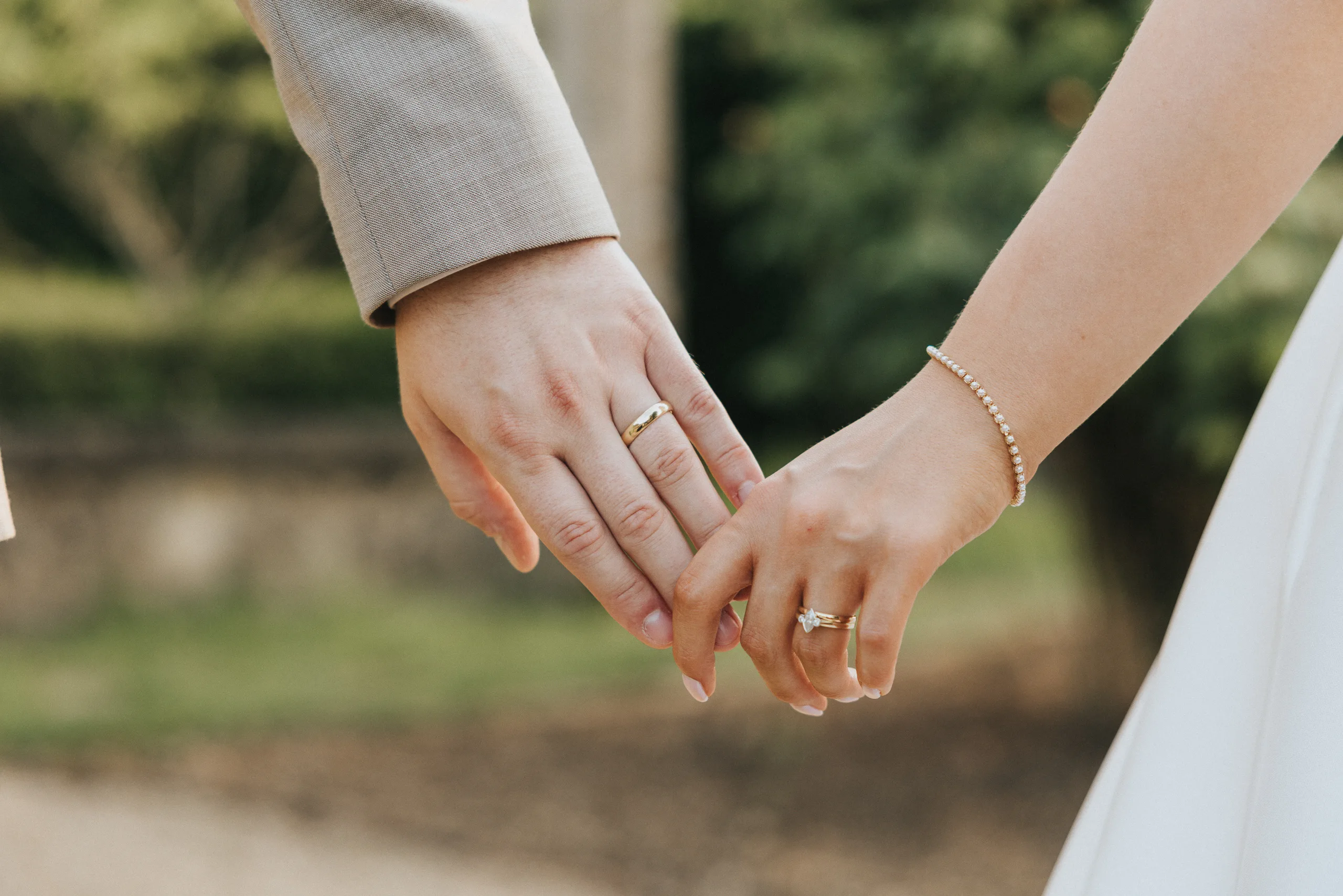 Close-up of two people holding hands outdoors at a Harlaxton Manor Wedding. Both wear rings on their ring fingers—one in a beige suit, the other in a white dress with a delicate bracelet. Blurred greenery and a stone wall complete the scene. © Aimee Lince Photography, Lincolnshire photographer