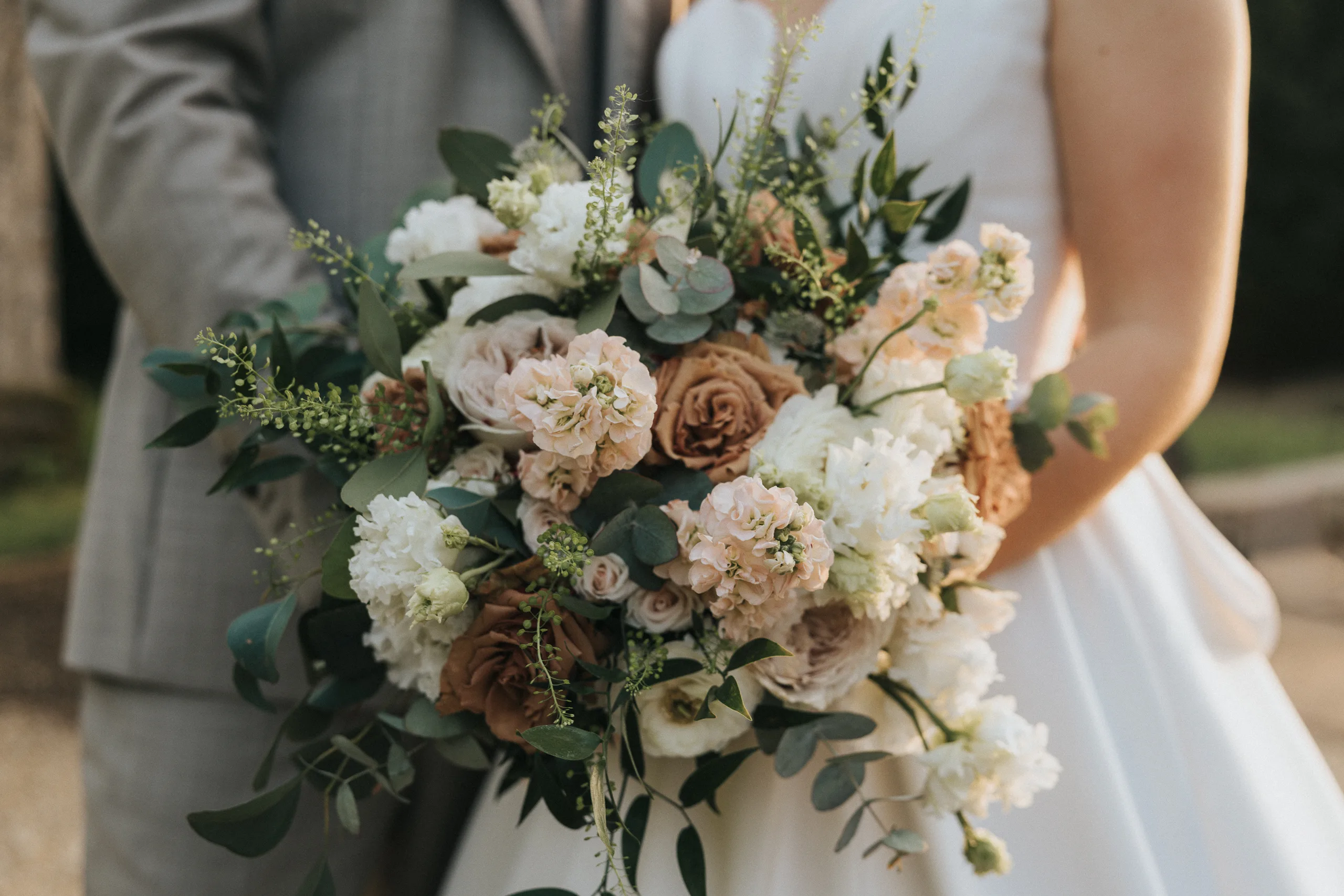 A bride in a white dress holds a large bouquet of white, blush, and tan roses with greenery and delicate filler flowers. Beside her stands another in a light grey suit. The softly blurred background hints at a romantic Harlaxton Manor Wedding. © Aimee Lince Photography, Lincolnshire photographer