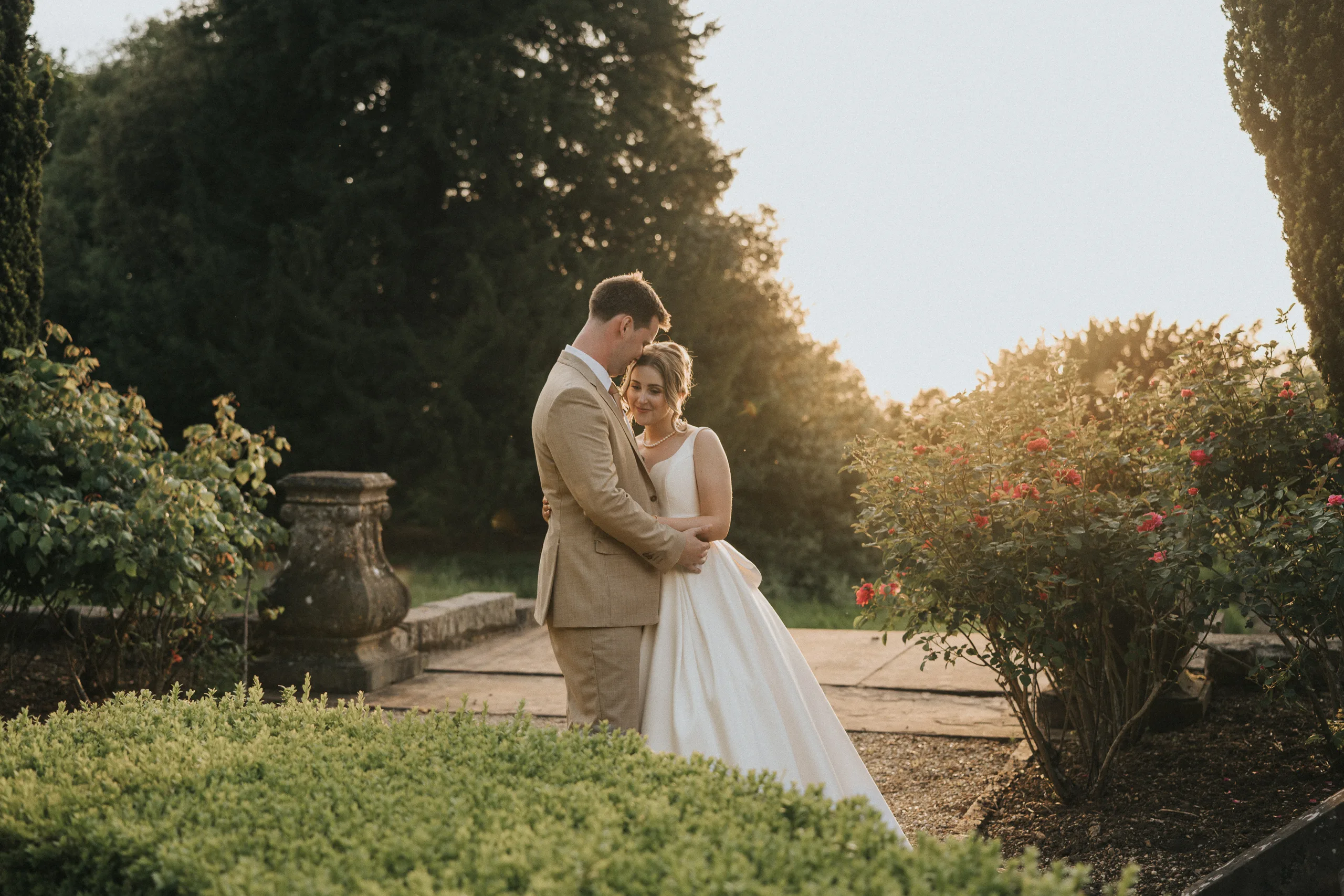 A bride in a white dress and a groom in a tan suit embrace and smile at each other in a sunlit garden during their Harlaxton Manor Wedding, surrounded by green bushes, blooming roses, tall trees, and soft evening light. © Aimee Lince Photography, Lincolnshire photographer