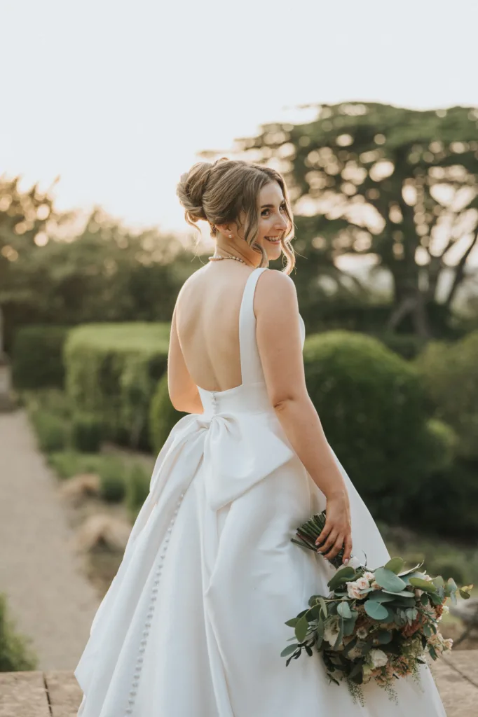A bride in a white, backless gown with a large bow stands outdoors at her Harlaxton Manor wedding, holding a bouquet of greenery and white flowers. She turns slightly, smiling over her shoulder as sunlight filters through lush trees in the background. © Aimee Lince Photography, Lincolnshire photographer