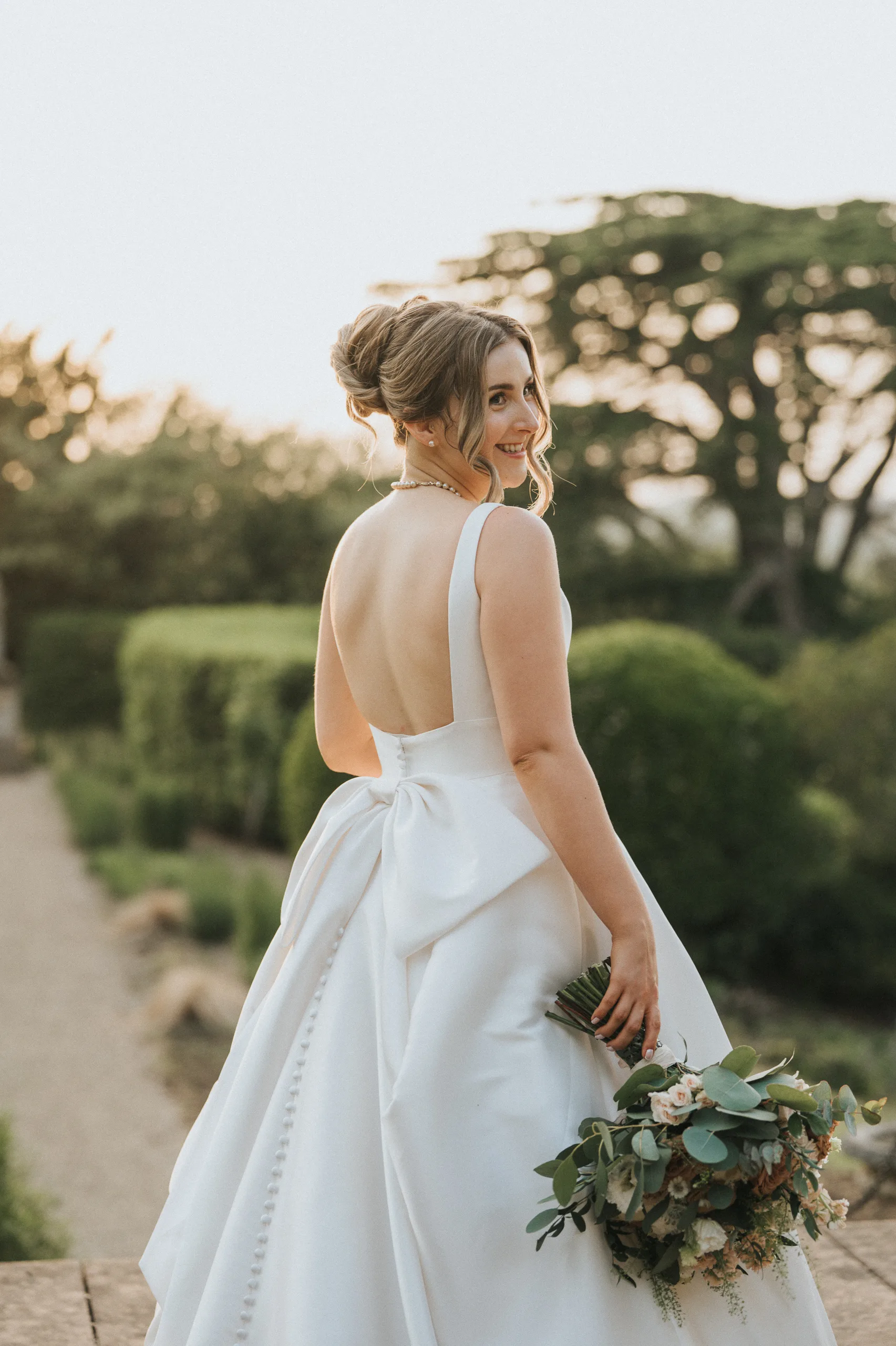 A bride in a white, backless gown with a large bow stands outdoors at her Harlaxton Manor wedding, holding a bouquet of greenery and white flowers. She turns slightly, smiling over her shoulder as sunlight filters through lush trees in the background. © Aimee Lince Photography, Lincolnshire photographer