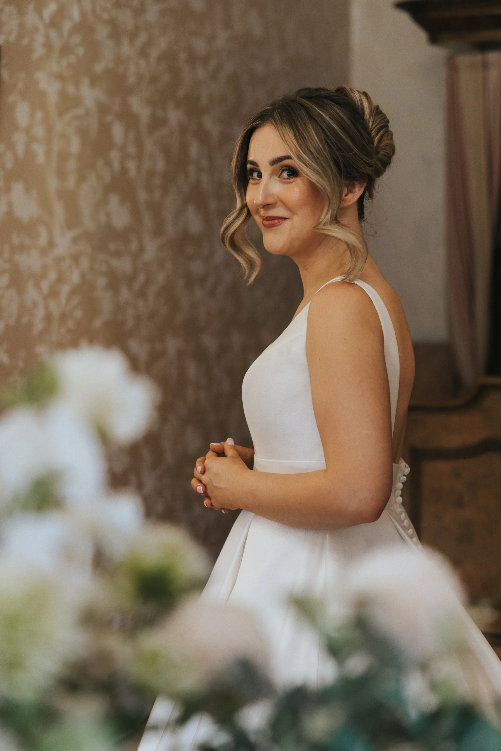 A smiling bride with light brown hair in an elegant updo wears a sleeveless, white satin wedding dress with a low back. She stands indoors at her Harlaxton Manor Wedding, hands clasped amid softly blurred white flowers and patterned wallpaper. © Aimee Lince Photography, Lincolnshire photographer