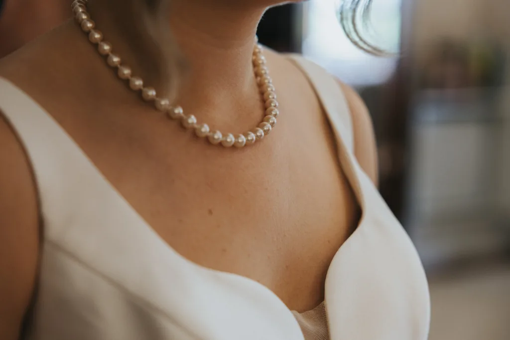 A close-up of a woman at a Harlaxton Manor Wedding, wearing a white sleeveless dress and single-strand pearl necklace. Her skin is softly lit and a loose strand of hair falls near her neck, with the blurred background highlighting her neckline and jewellery. © Aimee Lince Photography, Lincolnshire photographer