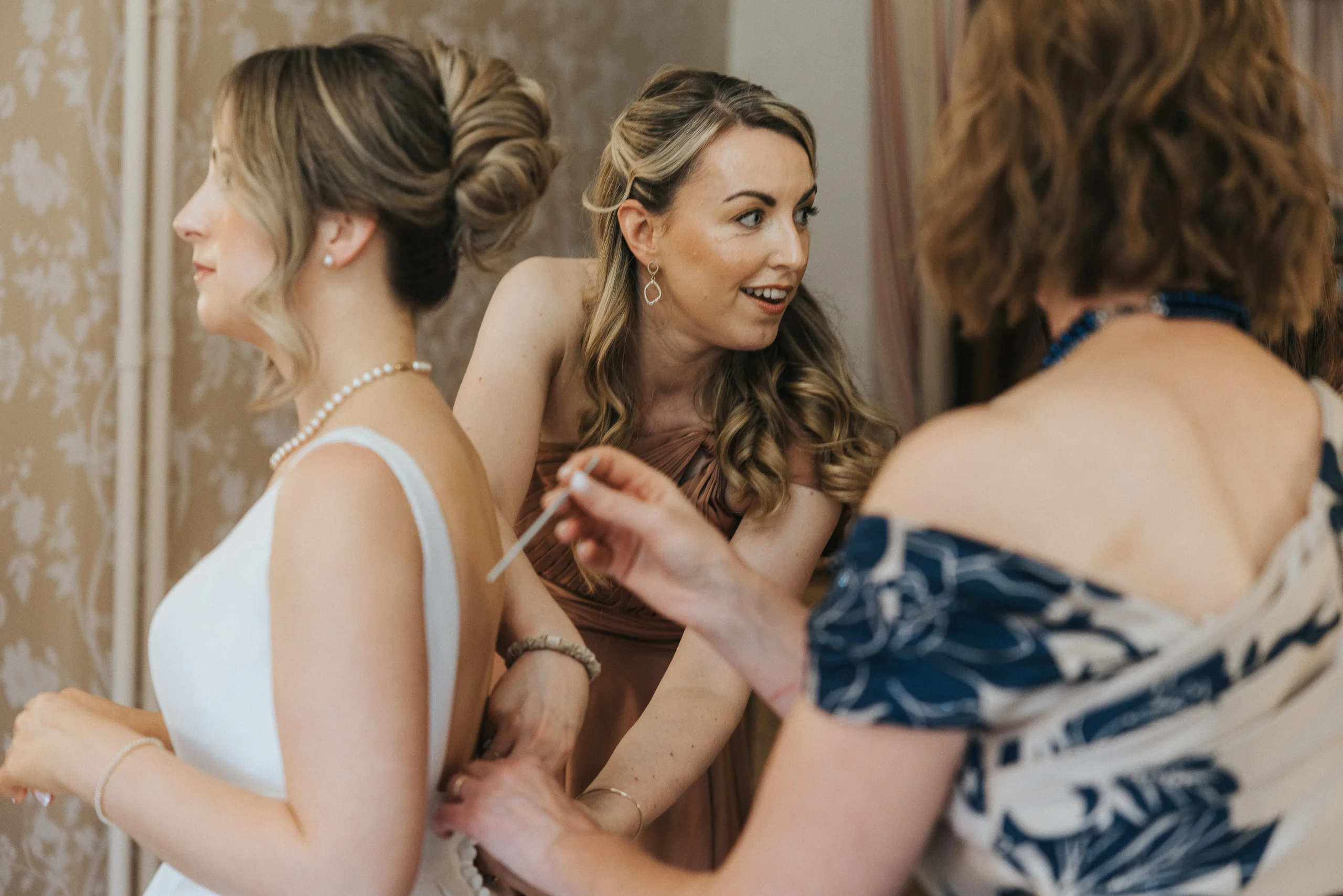 Three women prepare for a Harlaxton Manor Wedding. One, in a white dress, has her back fastened by a woman in florals, whilst another in peach smiles and assists. They stand indoors against light patterned wallpaper. © Aimee Lince Photography, Lincolnshire photographer