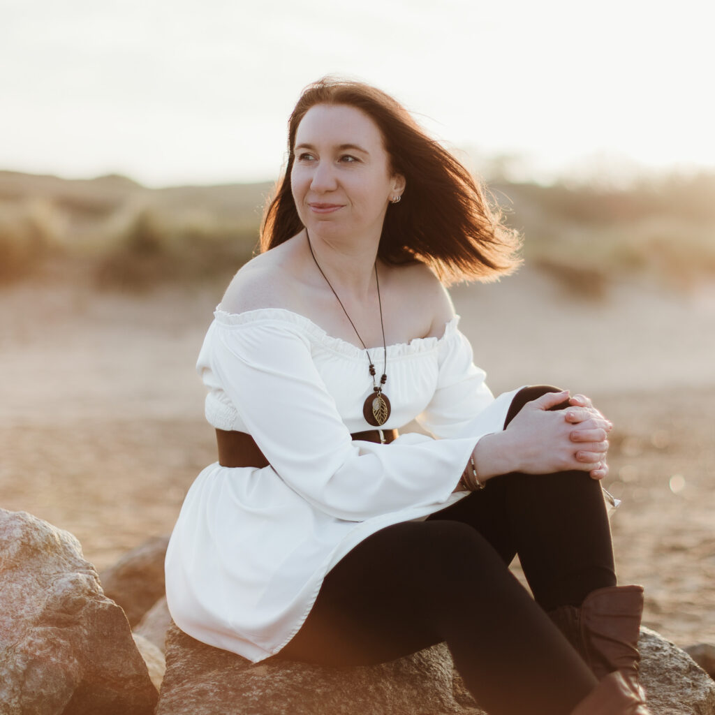 A woman with shoulder-length brown hair sits on a large rock on a sandy beach at sunset. She wears an off-the-shoulder white top with a dark wide belt, black leggings, brown boots, and a pendant necklace. Her hair blows slightly in the wind as she looks towards the horizon. © Aimee Lince Photography