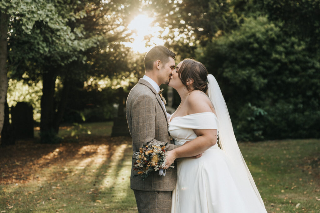 A bride in an off-the-shoulder white gown and veil kisses her groom, who wears a brown tweed suit and holds dried flowers in a sunlit park captured by a wedding photographer in Lincolnshire. Golden sunlight filters through the trees behind them. © Aimee Lince Photography