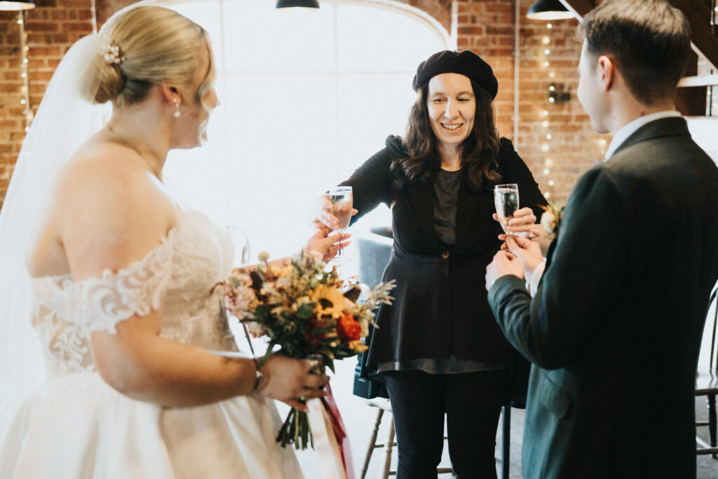 A bride in a white gown and a groom in a dark suit toast with a smiling woman in a black beret and blazer. Captured by a wedding photographer in Lincolnshire, they hold champagne flutes in a brick-walled room with fairy lights, the bride holding a vibrant autumn bouquet. © Aimee Lince Photography