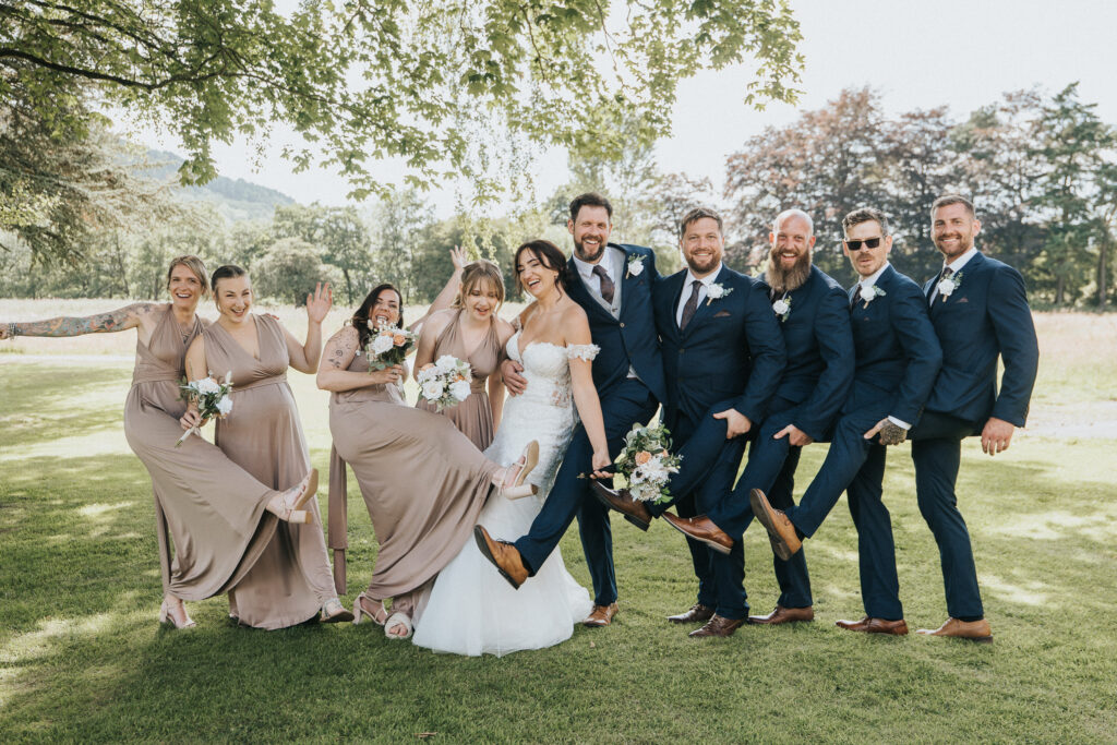 A wedding party in a Lincolnshire field shows off their shoes, with the bride in a white lace dress flanked by bridesmaids in tan dresses and groomsmen in navy blue suits playfully lifting their legs. © Aimee Lince Photography
