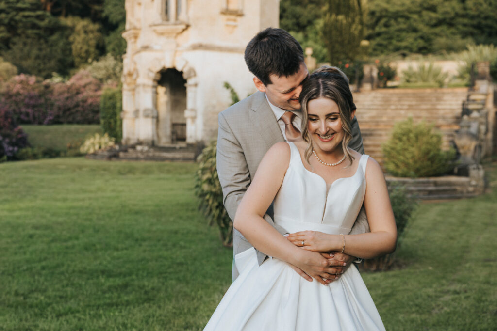 A bride in a white wedding dress and pearl necklace smiles as a man in a tan suit embraces her from behind in a Lincolnshire garden. They stand before an historic stone building with arched doorways, captured by Aimee Lince. © Aimee Lince Photography