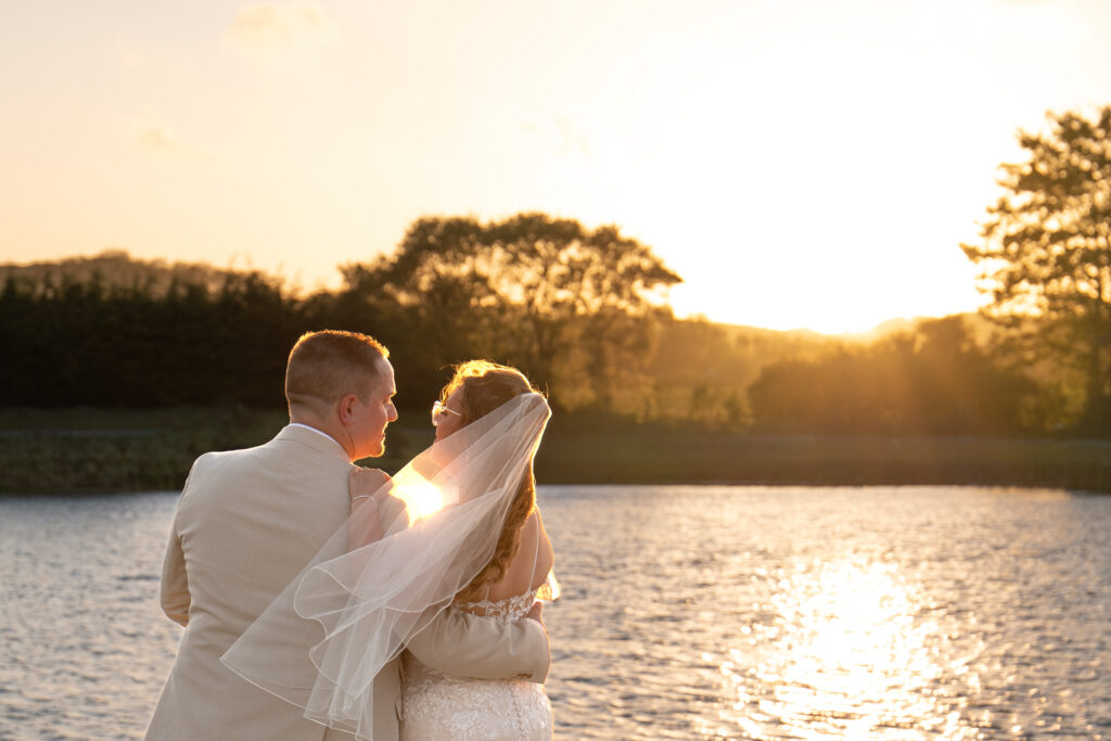 A bride and groom embrace whilst looking towards each other against a lake and sunset, a scene captured by a wedding photographer in Lincolnshire. The groom wears a tan suit, and the bride wears a white lace wedding dress with a veil, illuminated by golden light reflecting off the water. © Aimee Lince Photography