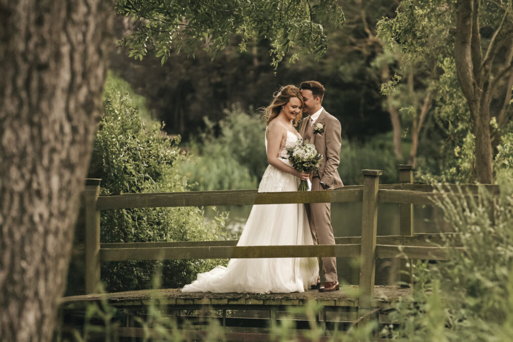 A bride in a white lace-bodice wedding dress and a groom in a tan suit stand close together on a wooden footbridge. They are surrounded by lush green trees in a Lincolnshire park, with the groom gently leaning in towards the bride as captured by a wedding photographer. © Aimee Lince Photography