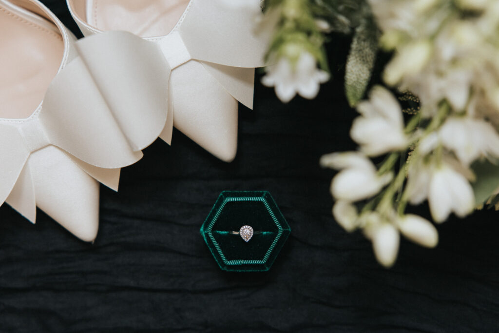 A pear-shaped diamond engagement ring rests in a green velvet hexagonal box on black fabric, captured by a Lincolnshire wedding photographer alongside white satin bow heels and a cluster of flowers. © Aimee Lince Photography