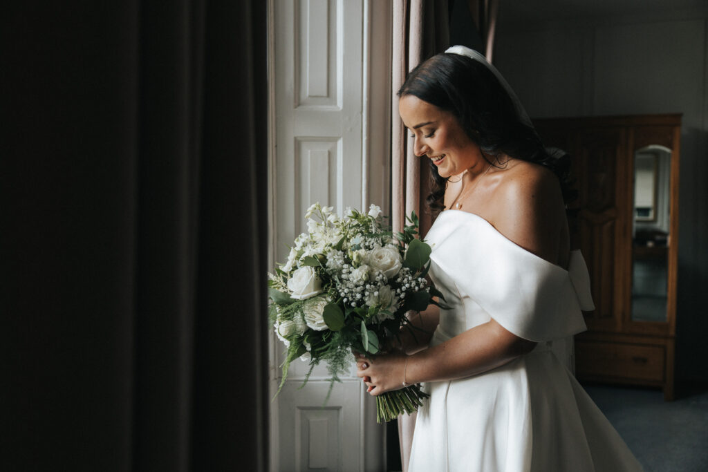A bride in a white off-the-shoulder gown smiles down at her bouquet of roses and greenery. Standing indoors near curtained windows, her veiled hair and moody atmosphere are captured by a wedding photographer in Lincolnshire. © Aimee Lince Photography