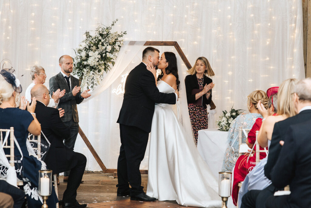 A bride and groom kiss at the altar in this Lincolnshire wedding photography shot as guests applaud. They stand before a wooden hexagonal arch draped with white fabric and glowing fairy lights, while a woman in a black blazer and patterned dress stands to the side. © Aimee Lince Photography