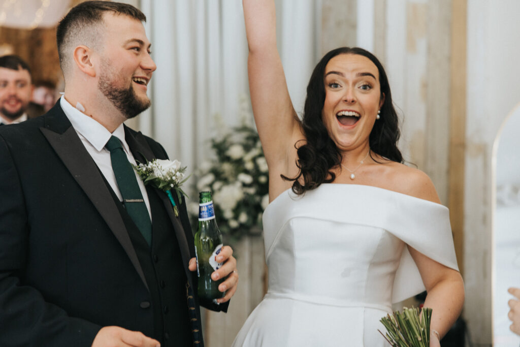A bride in a white off-the-shoulder gown cheers with her bouquet raised, whilst the groom laughs with a beer in his hand. Captured by a Lincolnshire wedding photographer, the formally dressed couple celebrate at their reception in a dark suit with a green tie. © Aimee Lince Photography