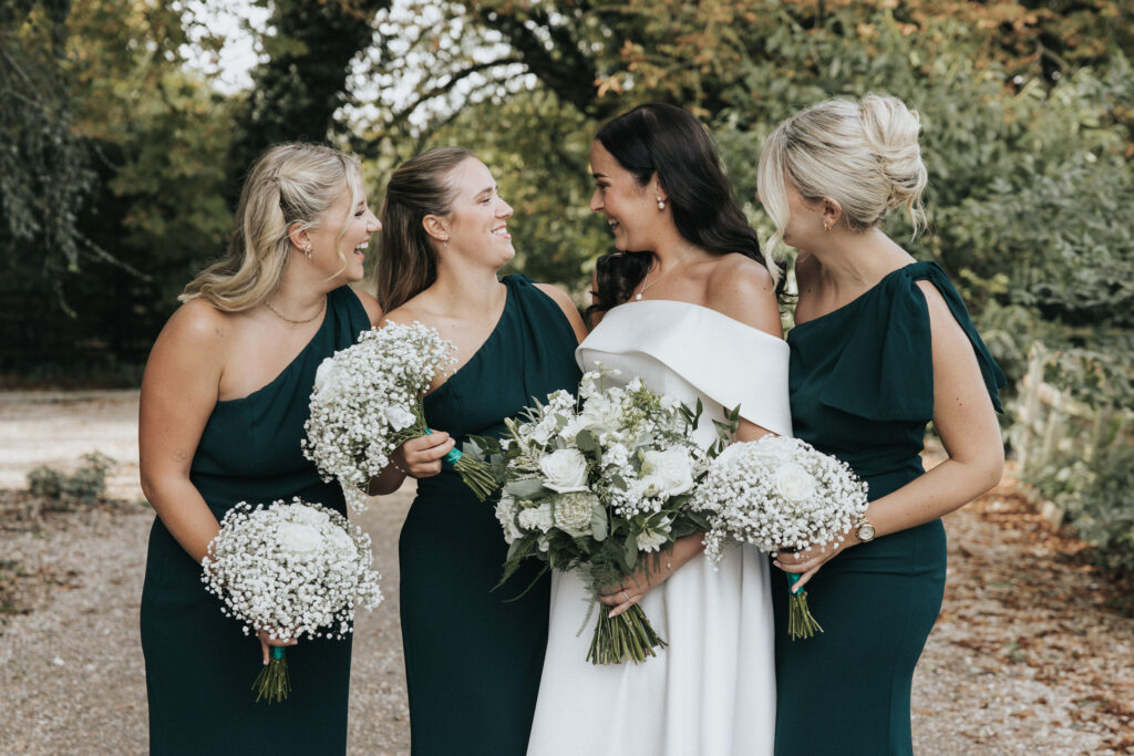A smiling bride in an off-the-shoulder white gown stands with three bridesmaids in matching teal dresses. Each woman holds a bouquet of roses and gypsophila on a tree-lined gravel path in this wedding photograph from Lincolnshire. © Aimee Lince Photography