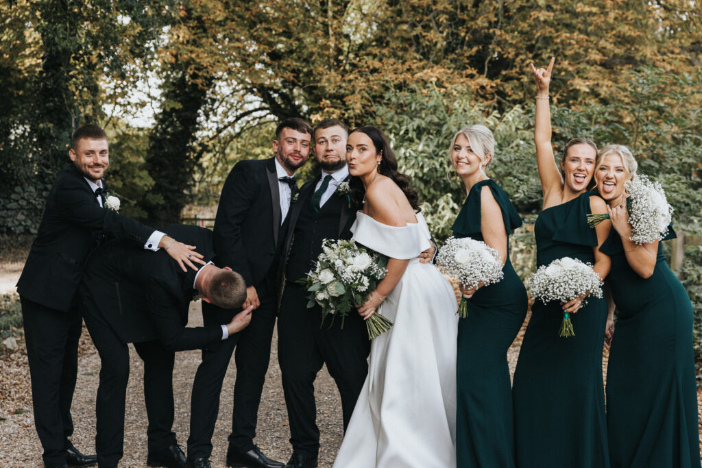 A wedding party poses outdoors with autumn trees in the background. A groom and his groomsmen in black suits surround a bride in an off-the-shoulder white gown, while three bridesmaids in dark green dresses hold bouquets; one bridesmaid makes a rock-on gesture in this Lincolnshire wedding photography. © Aimee Lince Photography