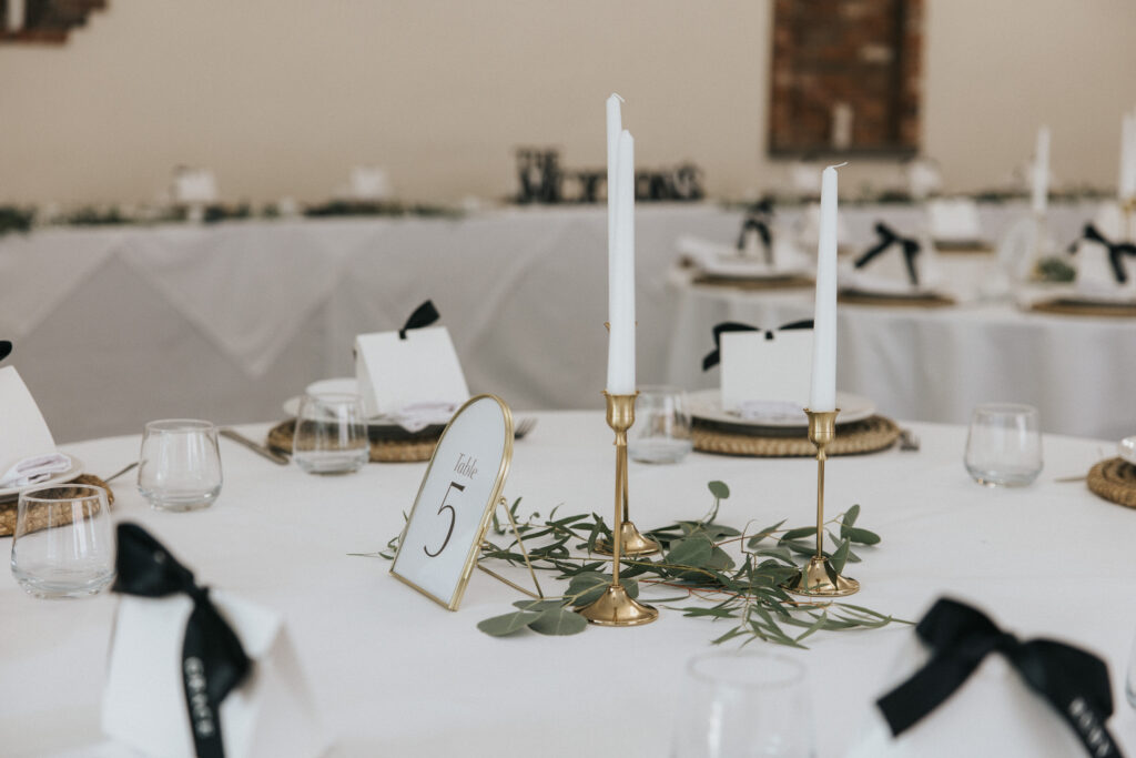 A round reception table features a gold-framed “Table 5” sign, two tall white tapers in brass holders, and a sprig of greenery, captured in a Lincolnshire wedding photography style. Background tables with white tablecloths and black-ribboned name cards are softly blurred. © Aimee Lince Photography