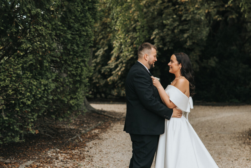 A bride and groom gaze lovingly at each other on a tree-lined gravel path in this Lincolnshire wedding photography. The groom wears a black suit, and the bride wears an off-the-shoulder white wedding dress with a bow detail on the back. © Aimee Lince Photography
