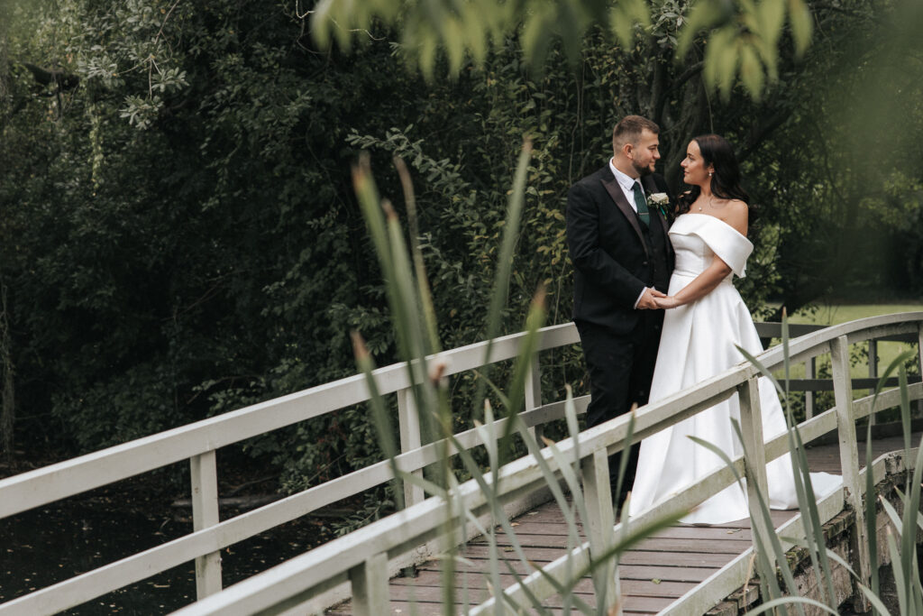 A bride and groom hold hands while facing each other on a wooden footbridge. The groom wears a black suit with a green tie, while the bride wears an off-the-shoulder white wedding dress, surrounded by the lush foliage captured by a wedding photographer in Lincolnshire. © Aimee Lince Photography