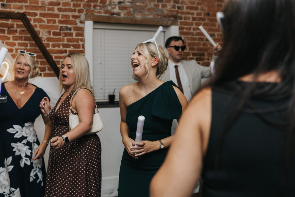 A smiling woman in a dark green one-shoulder dress holds a glowing foam baton at a wedding. Beside her, guests in formal wear dance in front of a rustic brick wall, a joyful moment captured by a Lincolnshire wedding photographer. © Aimee Lince Photography