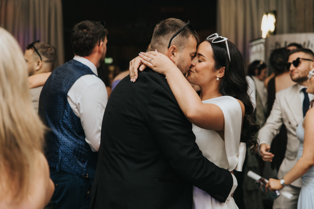 A bride in a white dress with heart-shaped sunglasses and a groom in a black suit share a kiss on a dance floor surrounded by wedding guests. This shot, captured by a Lincolnshire wedding photographer, shows guests in formal attire mingling in the blurred background. © Aimee Lince Photography