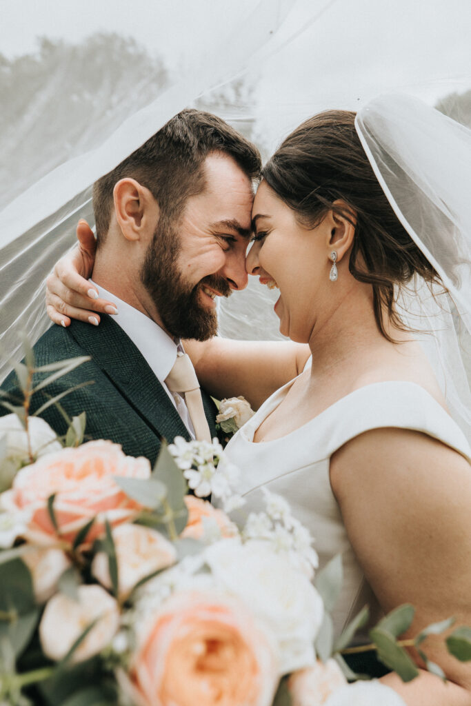 A bride and groom touch foreheads and smile under a sheer veil, captured by a wedding photographer in Lincolnshire. The groom wears a dark suit with a light tie, whilst the bride wears a white gown and holds a bouquet of soft-coloured flowers in the foreground. © Aimee Lince Photography