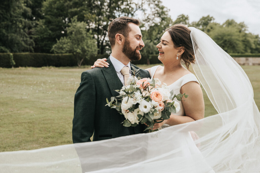 A bride and groom share a laugh in a grassy Lincolnshire field. The bride wears a white wedding dress and veil, while the groom wears a dark green suit. She holds a bouquet of white and peach flowers as her long veil blows across the foreground in this wedding photographer image. © Aimee Lince Photography
