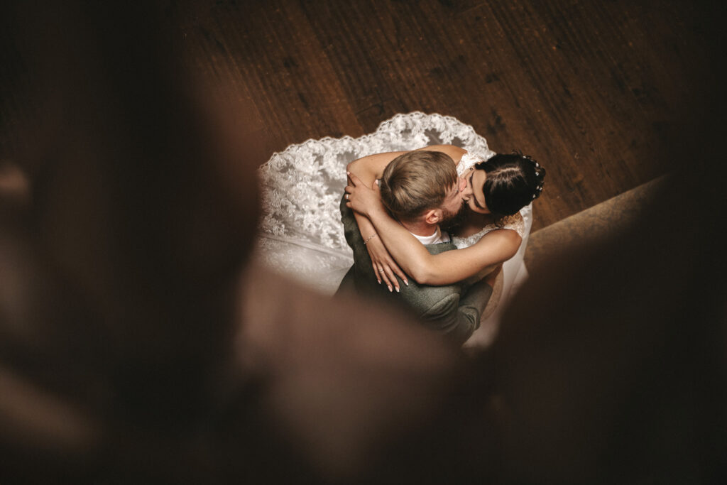 A bride and groom embrace and kiss while dancing on a dark wooden floor, captured by a wedding photographer in Lincolnshire. The bride wears a white lace wedding dress with a long train, whilst the groom is in a dark suit, framed by blurred foreground elements in a soft, intimate scene. © Aimee Lince Photography
