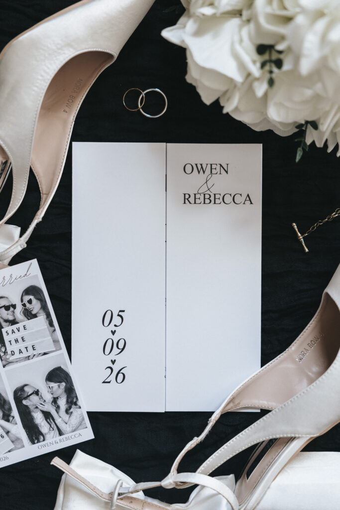 A wedding invitation for Owen and Rebecca is centred on a black surface alongside two white high heels, wedding bands, and a photo strip. The stationery, styled for a documentation home, features the date 05.09.26 and the couple’s names in elegant text near a white floral arrangement. © Aimee Lince Photography