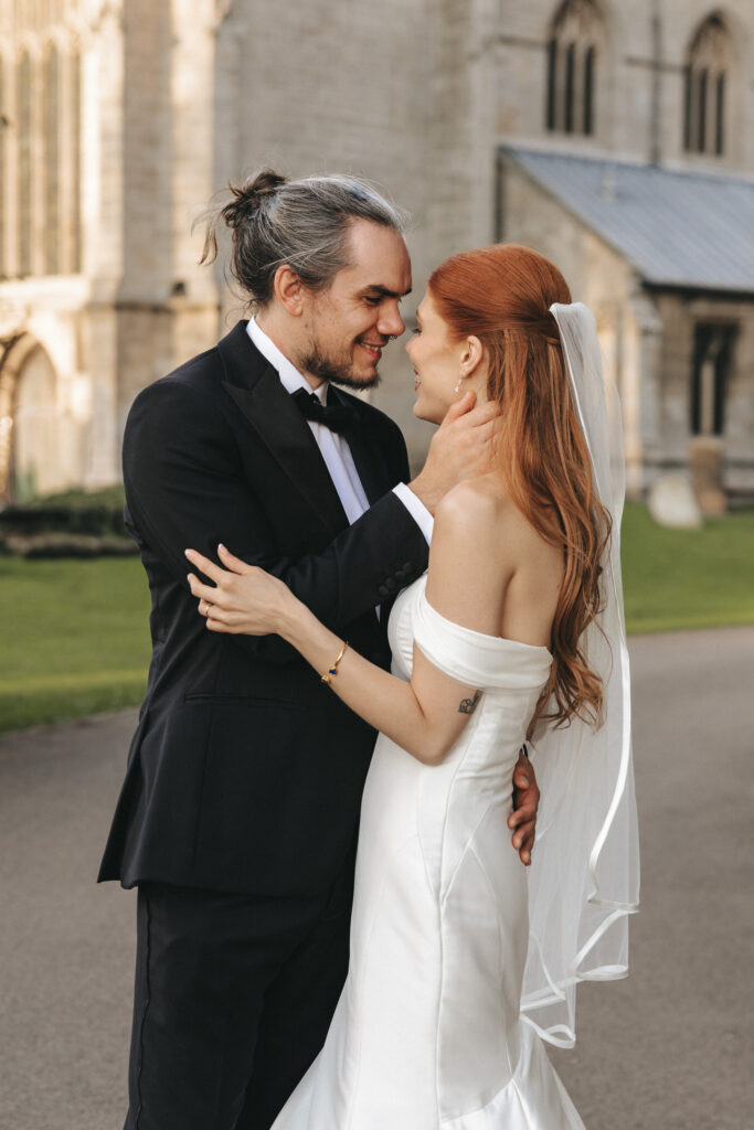 A bride and groom gaze lovingly at each other on a path before a stone church at The Beonna in Lincolnshire. The groom wears a black tuxedo with a topknot, while the bride wears an off-the-shoulder white gown and a veil clipped to her long red hair. © Aimee Lince Photography