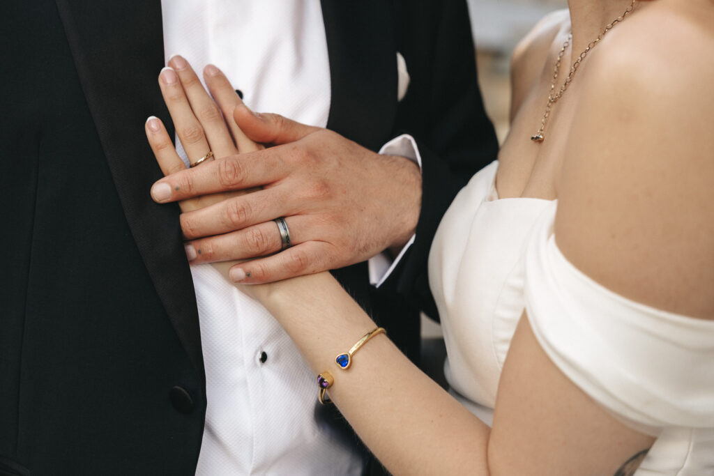 A bride in a white off-the-shoulder dress rests her hand, adorned with a gold bracelet, on a groom’s tuxedo-clad chest to showcase their wedding rings. © Aimee Lince Photography