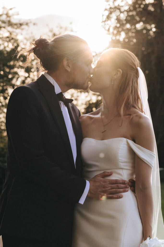 A bride in a white off-the-shoulder gown and a groom in a black tuxedo kiss outdoors at sunset. The bright sun creates a warm lens flare, casting the couple in silhouette at The Beonna in Lincolnshire. © Aimee Lince Photography