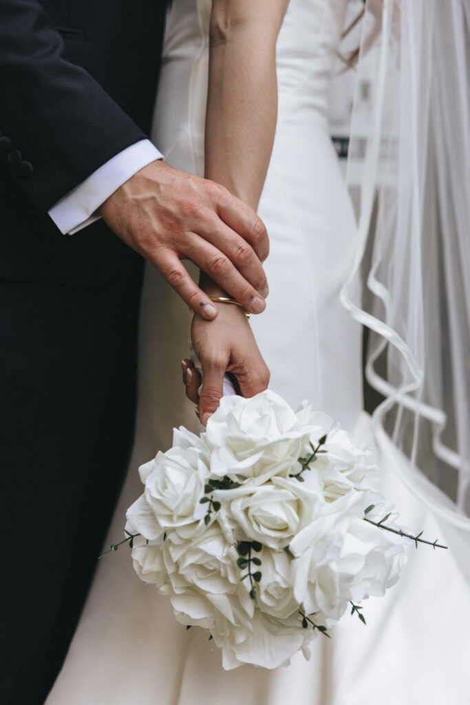 A bride in a simple white gown and veil holds hands with a groom in a black tuxedo. She carries a bouquet of white roses, and a delicate gold band is visible on her finger. © Aimee Lince Photography
