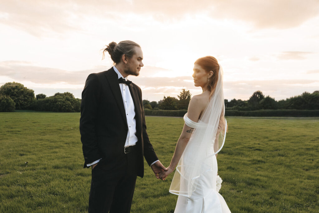 A bride in a white strapless gown and veil holding hands with a groom in a black tuxedo stand in a grassy field. They gaze at each other in profile against a golden sunset at The Beonna in Lincolnshire, with a line of dark trees bordering the open meadow. © Aimee Lince Photography